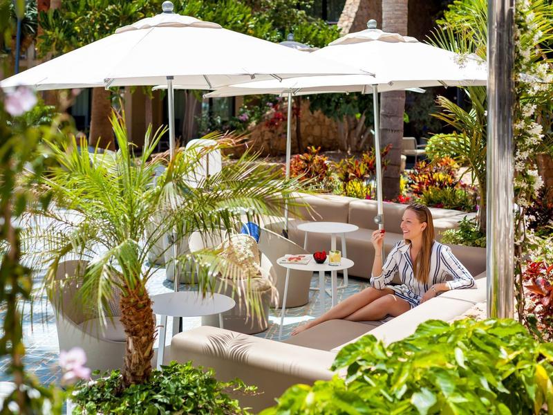 Femme se détend sur une chaise longue près de la piscine sous des parasols dans un jardin tropical.