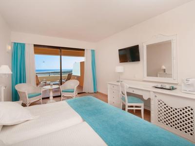 Hell eingerichtetes Hotelzimmer mit zwei Betten, blauem Vorhang und Balkon mit Meerblick.