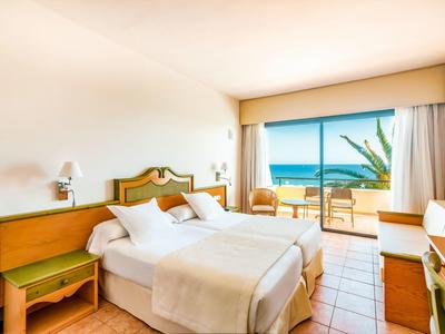 Helles Hotelzimmer mit Doppelbett, Balkon und Meerblick, hellen Wänden und Holzmöbeln.