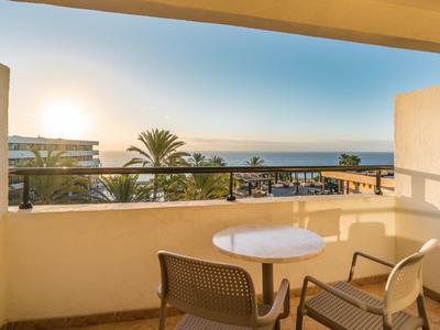 Balkon mit zwei Stühlen und Tisch, Blick auf Palmen, Gebäude und Meer bei Sonnenuntergang.