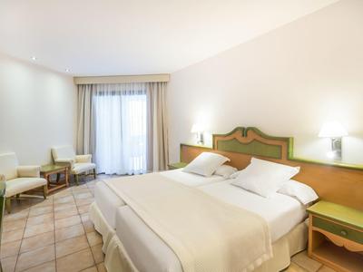 Helles Schlafzimmer mit großem Bett, grünen Nachttischen, Sesseln und Fliesenboden.