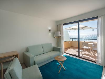 Helles Wohnzimmer mit blauem Teppich, Sofa, Sessel, kleiner Holztisch und Balkon mit Meerblick.
