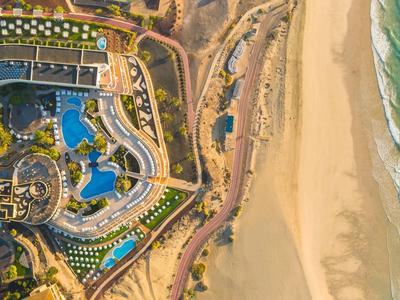 Luftaufnahme eines Resorts mit Pool, Strandliegen und breitem Sandstrand neben türkisfarbenem Meer.