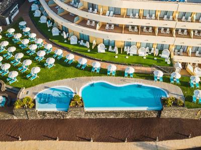 Hotelanlage mit rundem Pool, Liegestühlen auf grünem Rasen und mehrstöckigem Gebäude im Hintergrund.