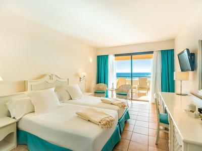 Helles Hotelzimmer mit großen Betten, türkisfarbenen Vorhängen und Blick auf den Meerblick.