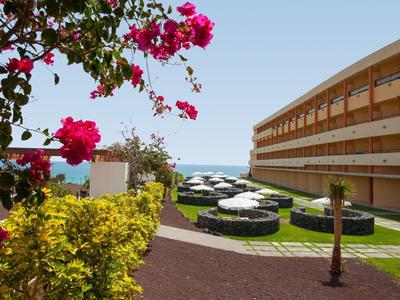 Blick auf Hotel mit Terrasse, Sonnenschirmen und Blumen im Vordergrund, unter blauem Himmel.