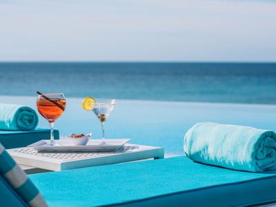 Luxuriöser Pool mit türkisfarbenen Handtüchern und Getränken, Meer im Hintergrund sichtbar.