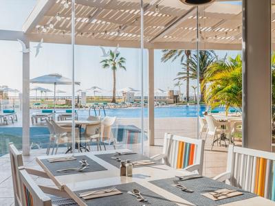 Terrassenrestaurant mit Holztischen, bunten Stühlen, Poolblick und Palmen bei Sonnenschein.