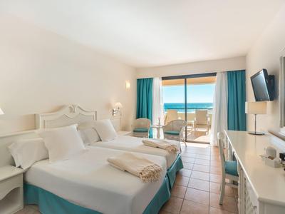 Helles Hotelzimmer mit zwei Betten, blauen Vorhängen und Meerblick vom Balkon.