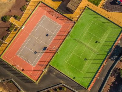 Zwei nebeneinander liegende Sportplätze, ein oranger Tennisplatz und ein grüner Fußballplatz, umgeben von Zäunen.