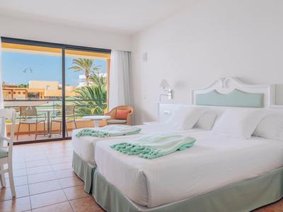 Helles, weiß eingerichtetes Schlafzimmer mit Doppelbett, Balkon und Blick auf Palmen und Gebäude.