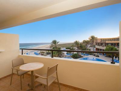 Balkon mit zwei Stühlen und Tisch, Blick auf Pool, Palmen und Meer am Horizont unter blauem Himmel.