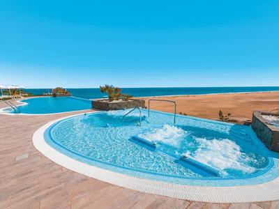 Mehrere blaue Swimmingpools mit Wasserdüsen und einem hellen Holzboden vor einem weiten Sandstrand.
