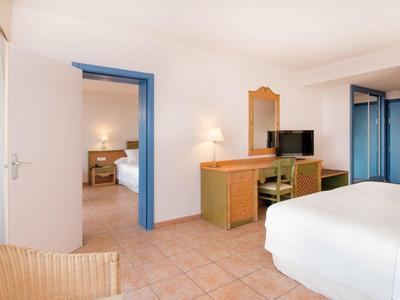 Helles Hotelzimmer mit Doppelbett, Holzmöbeln, Fliesenboden und Blick ins angrenzende Schlafzimmer.