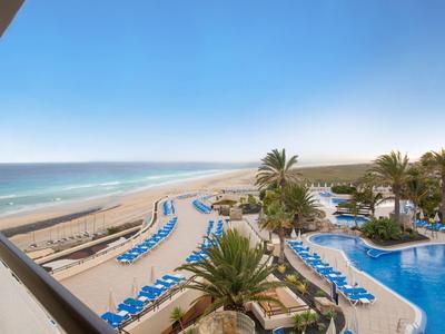 Strandhotel mit blauem Pool, vielen Sonnenliegen und Palmen unter klarem Himmel.