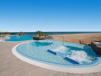 Moderner Pool mit Whirlpool, blauem Wasser und weiten Sandstrand im Hintergrund unter klarem Himmel.
