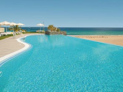 Großer blauer Pool mit Sonnenliegen am Strand und weitem Blick aufs Meer bei klarem Himmel.