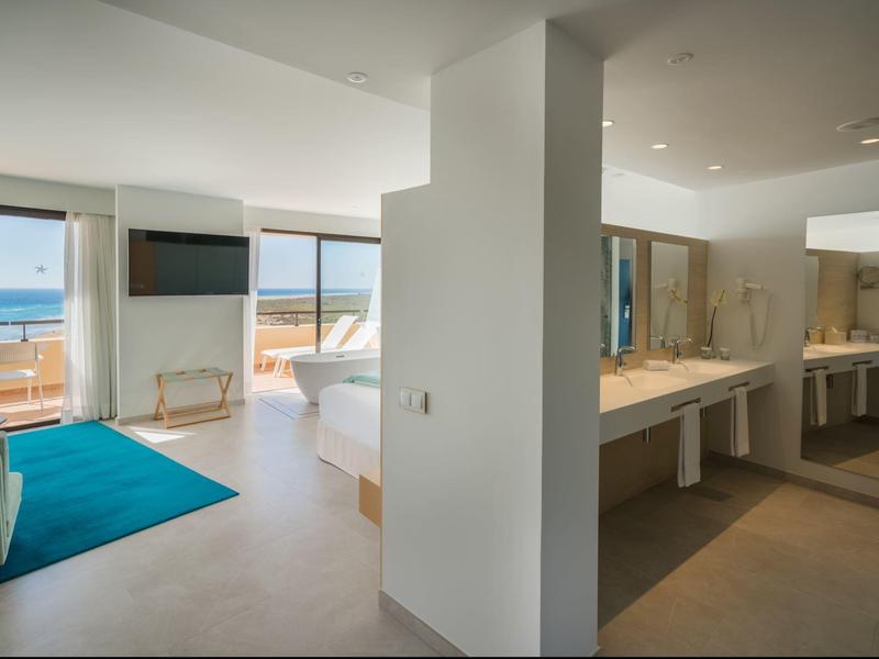 Przestronny pokój hotelowy z widokiem na ocean, niebieski dywan, łóżko, TV i dobrze oświetlona łazienka z podwójną umywalką.