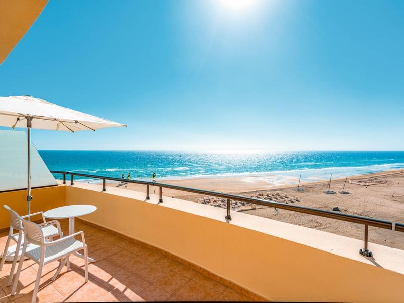 Słoneczny balkon z stołem, krzesłami, parasolem, widokiem na piaszczystą plażę i ocean pod czystym niebem.