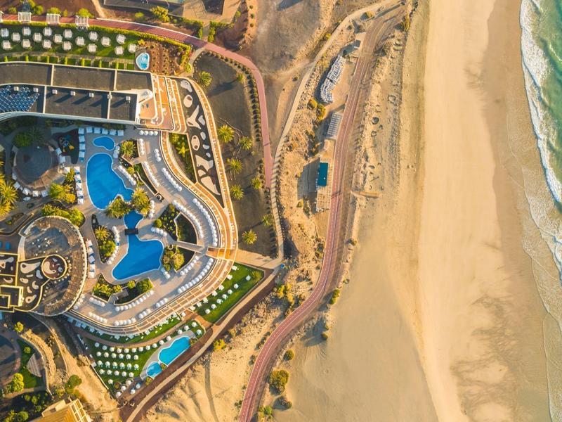 Widok z lotu ptaka na nadmorski ośrodek z basenami, budynkami i piaszczystą plażą nad morzem.