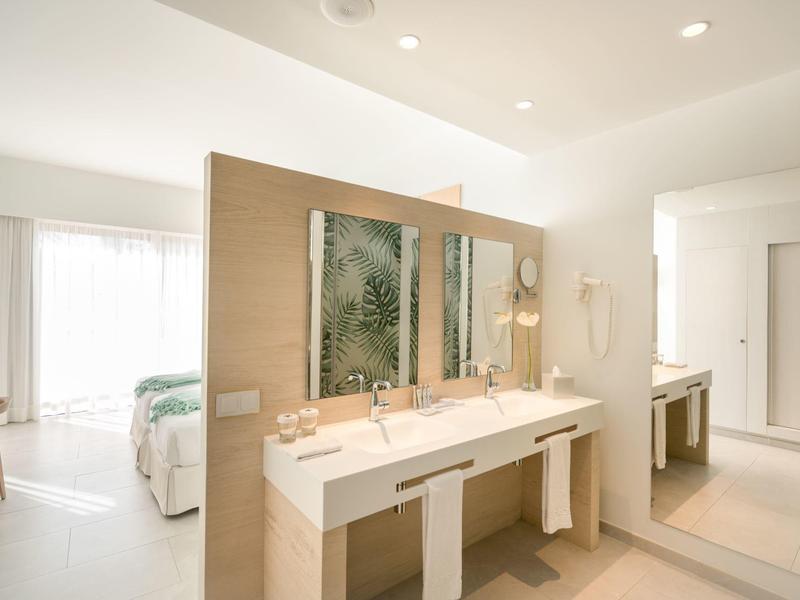 Jasna łazienka hotelowa z podwójną umywalką, dużym lustrem i widocznym łóżkiem w tle.