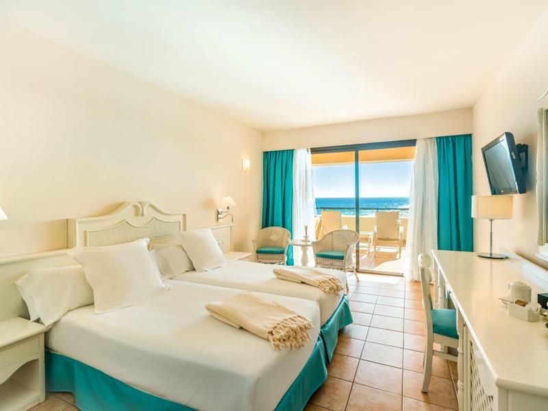 Jasny pokój hotelowy z podwójnym łóżkiem, balkonem i widokiem na morze przez przeszklone drzwi.