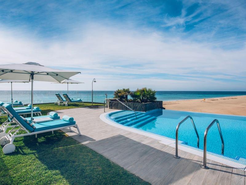 Leżaki przy basenie pod parasolami z widokiem na ocean i piaszczystą plażę.