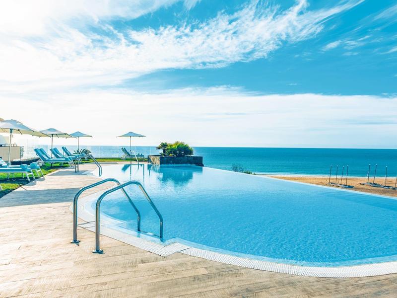 Basen typu infinity z widokiem na morze, leżaki i parasole w słoneczny dzień.