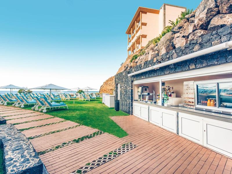 Bar na świeżym powietrzu z drewnianym tarasem i leżakami obok skalistego wzgórza pod czystym niebem.