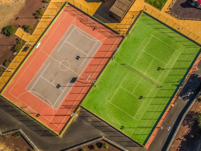 Widok z lotu ptaka na dwa przylegające boiska sportowe, jedno czerwone, drugie zielone, otoczone ogrodzeniami.