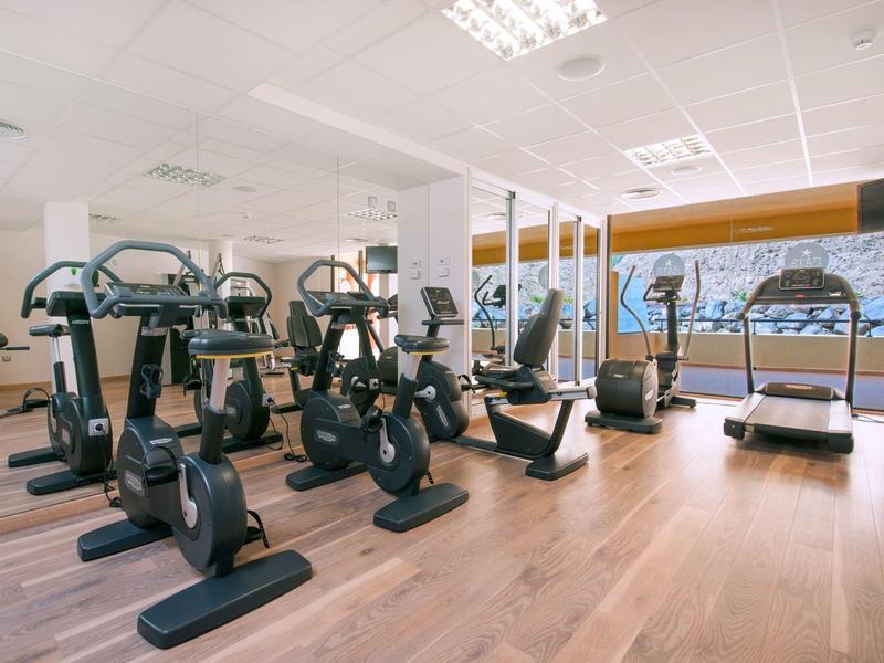 Jasna siłownia z urządzeniami cardio i drewnianą podłogą, duże okna z widokiem na zewnątrz.