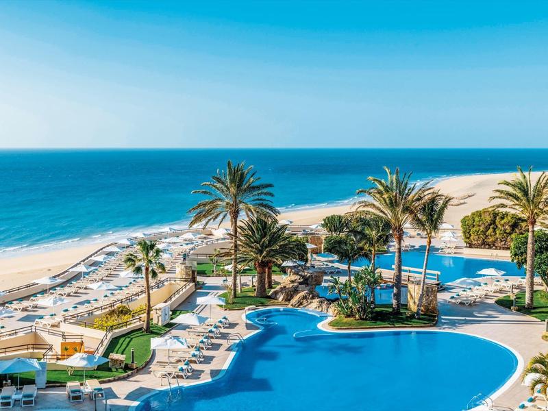 Ośrodek z dużym basenem, palmami, leżakami i piaszczystą plażą z widokiem na morze.