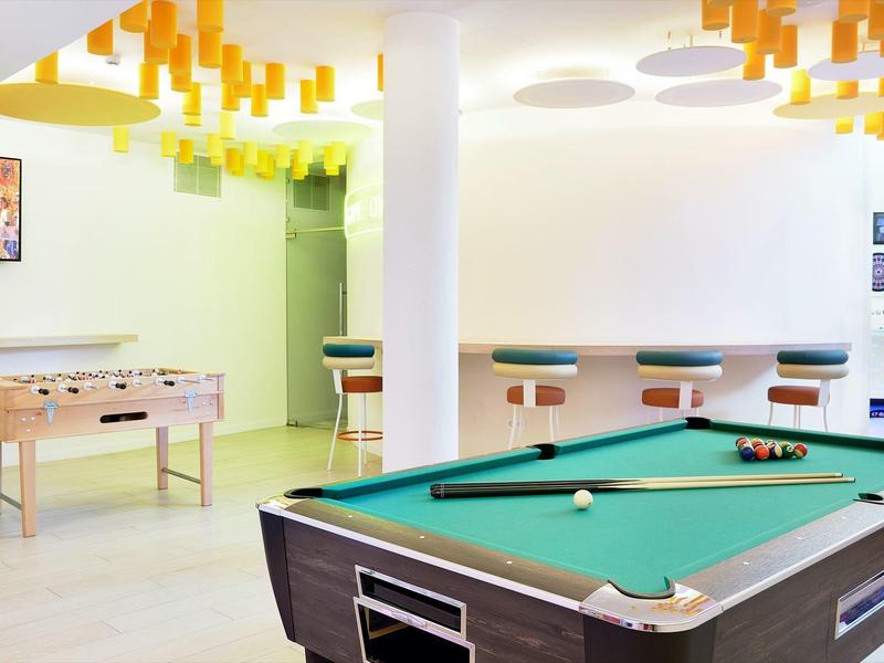 Jasny pokój gier z stołem bilardowym, piłkarzykami, stołkami barowymi i żółtymi lampami.