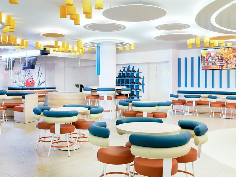 Nowoczesna lobby hotelowa z okrągłymi stołami, turkusowymi i brązowymi krzesłami oraz jasnymi okrągłymi światłami sufitowymi.