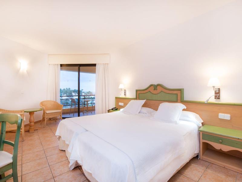 Jasny pokój hotelowy z podwójnym łóżkiem, zielonym drewnianym meblami i balkonem z widokiem na morze.