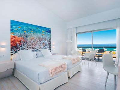 Helles Schlafzimmer mit Doppelbett, Meeresblick durch große Fenster, weiße Möbel und bunte Wandkunst.