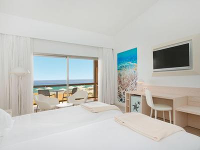Helles Hotelzimmer mit Doppelbett, Schreibtisch, Wandbild und Balkon mit Meerblick.