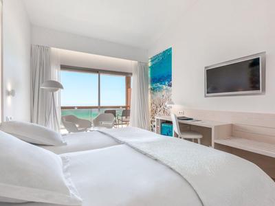 Helles Hotelzimmer mit Doppelbett, Flachbildfernseher, Schreibtisch und Balkon mit Meerblick.