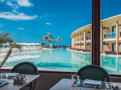 Blick aus Restaurant mit gedecktem Tisch auf großen blauen Pool und modernes Hotel bei sonnigem Himmel.