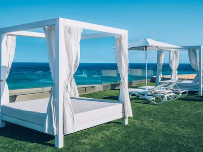 Weiße Lounge-Betten mit Vorhängen stehen auf grünem Rasen am blauen Meer unter klarem Himmel.