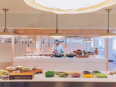 Moderne Buffet-Theke mit verschiedenen Speisen, Koch im Hintergrund, helle und freundliche Atmosphäre.