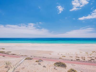 Breiter Sandstrand mit blauem Meer und teilweise bewölktem Himmel, Pfad im Vordergrund.