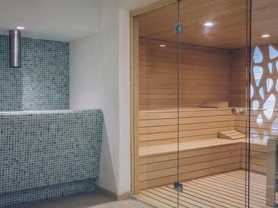Moderne Sauna mit Holzinnenraum und Glaswand, daneben Mosaikfliesen-Wand in Grau- und Blautönen.