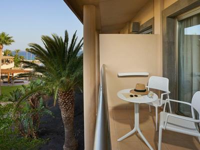 Balcone con tavolo bianco, due sedie e un cappello, vista su palme e area dell'hotel.