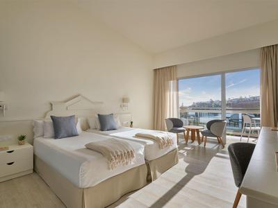 Camera d'hotel luminosa con due letti singoli, area salotto e vista sul mare attraverso grandi finestre.