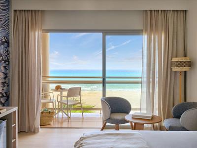 Camera d'hotel con balcone, vista sulla spiaggia e sul mare, area salotto moderna e tende chiare