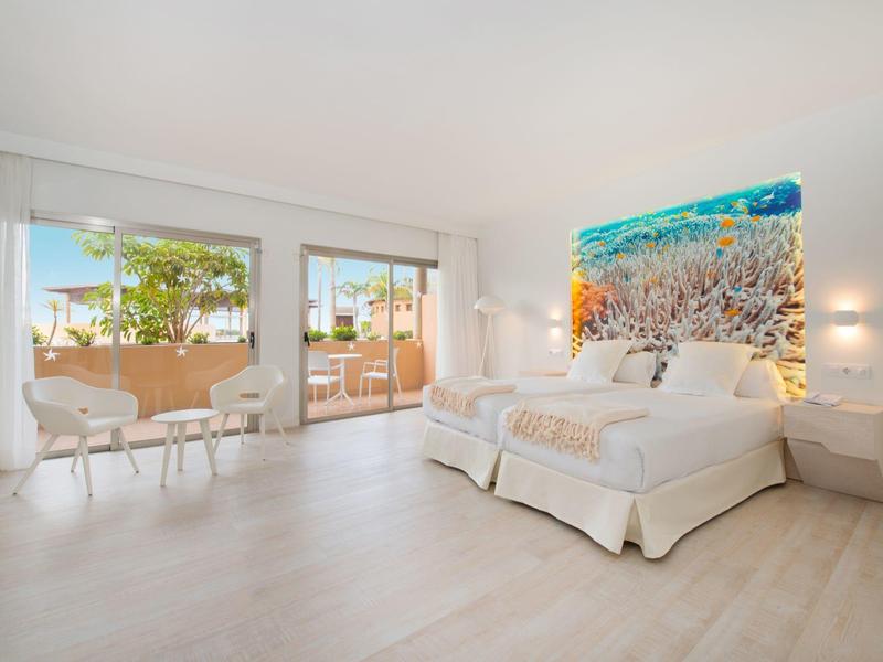 Camera d'hotel luminosa con letto matrimoniale, due sedie e grande balcone con vista sugli alberi.