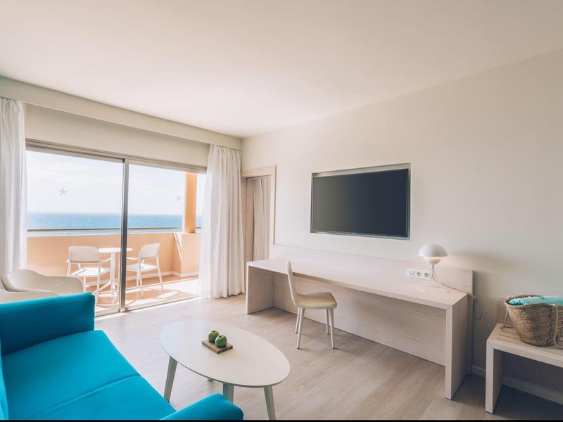 Camera d'hotel luminosa con divano blu, scrivania, sedia e balcone con vista sul mare.