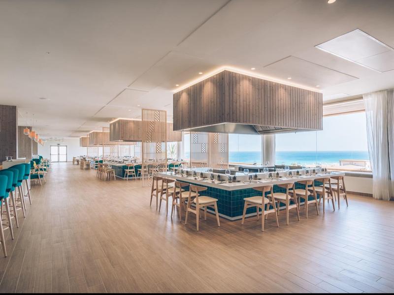 Ristorante luminoso e moderno con pavimento in legno, aree salotto e grandi finestre che lasciano entrare la luce del giorno.