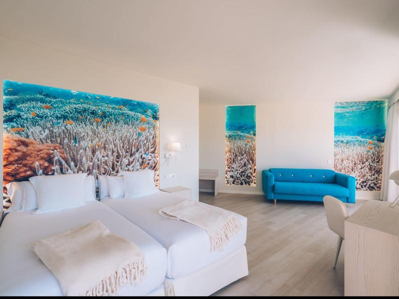 Camera d'hotel luminosa con due letti, un divano blu e decorazioni murali a tema oceano.